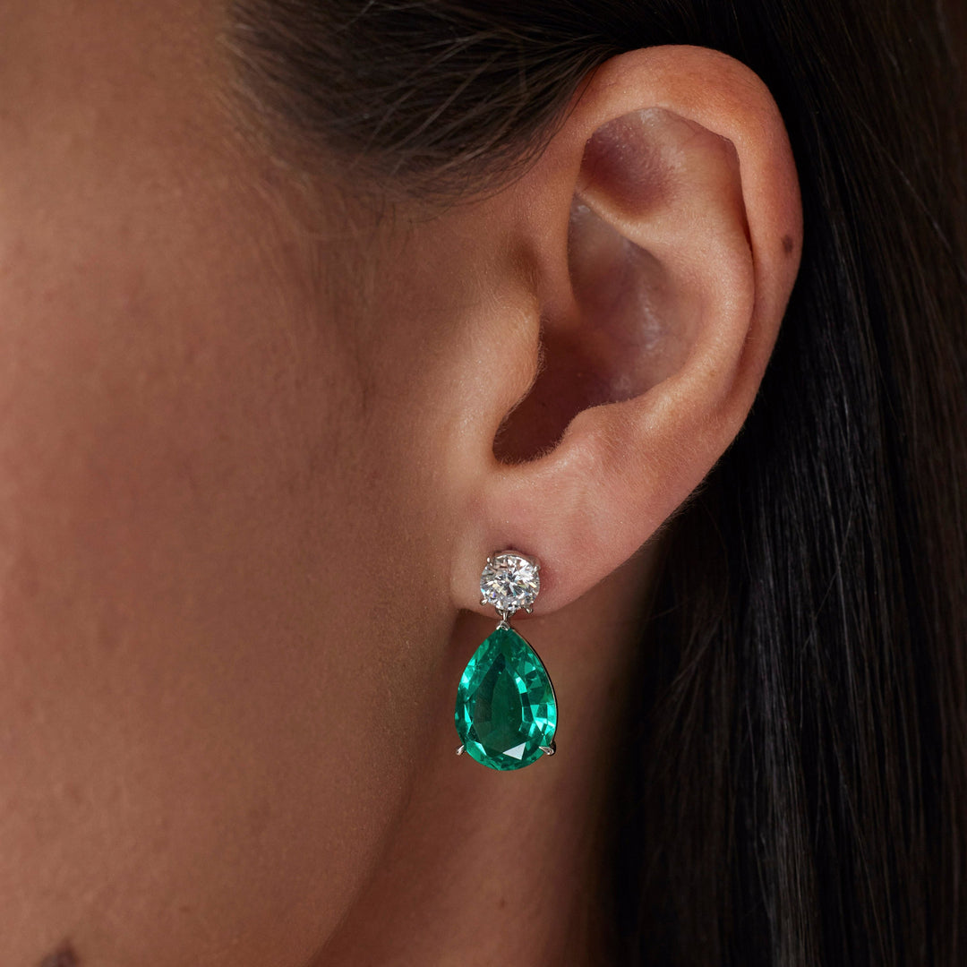 18k gold lab grown diamond and emerald earrings. Round brilliant and pear cut with tiger claw setting. On model.