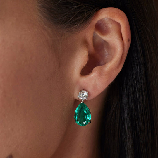 18k gold lab grown diamond and emerald earrings. Round brilliant and pear cut with tiger claw setting. On model.