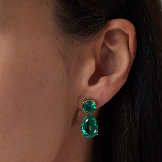 18k gold lab grown emerald earrings. pear and round brilliant cut emeralds. Designed in Zurich. On model.